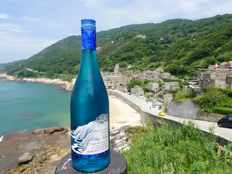 藍眼淚葡萄酒立在北竿芹壁海岸邊，後方是芹壁石屋聚落與海。